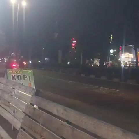 The video shows a nighttime street scene in Calukan RT 1 RW 5 Keboansikep, Indonesia. The camera is positioned behind a wooden bench, looking out at a road with traffic. In the foreground, the wooden slats of the bench are clearly visible.

On the left side of the road, there is a green sign with yellow lettering that reads "Aneka KOPI," suggesting a coffee shop or vendor. Further down the road, streetlights illuminate the scene. A few vehicles are visible, including a car with its brake lights on and what appears to be a motorcycle. In the distance, there are more lights from buildings and streetlights. One building has a prominent sign that appears to be a McDonald's restaurant. A large truck, possibly a semi-trailer truck, drives by in the middle of the road. The sky is dark, indicating it is nighttime, and the weather appears clear. The overall mood is calm and urban, with the ambient lights creating a somewhat subdued atmosphere. There are no people clearly discernible in the frame, though the presence of vehicles suggests activity.