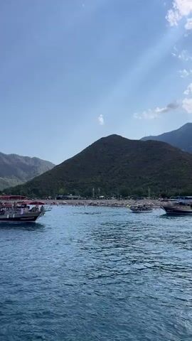 The video shows a body of water with several boats. In the background, there is a sandy beach crowded with people, and behind the beach, there are mountains covered with trees. The sky is blue with some clouds. The water appears calm, with gentle waves. Some of the boats are closer to the shore, possibly offering tours or transportation to the beach. The overall scene depicts a lively and scenic coastal area.