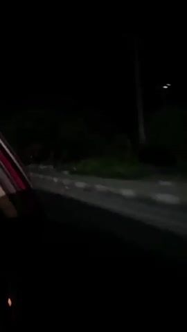 The video shows a view from a car driving along a dark road at night. The road has white concrete barriers on the right side. In the darkness, shapes of trees and houses are vaguely visible on the side of the road. The red and white trim of the car's interior is visible in the foreground.