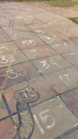 The video shows a young child wearing a pink outfit adorned with cartoon characters and a pink baseball cap, standing on a playground surface. The surface is painted with a large game board that resembles the game of Snakes and Ladders. Numbered squares, snakes, and a ladder are visible. The child is looking down and appears to be about to play the game. The background includes grass and what looks like park benches or seating. The video is shot from a low angle, focusing on the child and the game board.