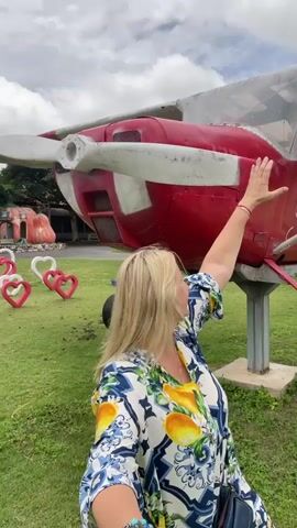 A blonde woman in a floral shirt is standing in a grassy field in front of a red and white airplane. She has her right hand on the airplane's propeller. She is looking at the camera and smiling. In the background, there are several heart-shaped decorations and some buildings. The sky is cloudy.