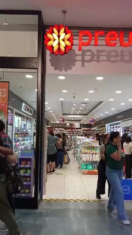 The video shows the entrance to a store called Preunic, which appears to be a pharmacy or beauty supply store, located in a shopping mall. The store is well-lit with bright interior lights. Several customers are inside, browsing the aisles. Display shelves are stocked with various products. Some customers are standing in a line towards the back of the store. People are walking past the store's entrance. There are advertisements on the left side of the entrance. A red and yellow floral-shaped light fixture hangs above the entrance.  The floor outside the store is tiled in gray.