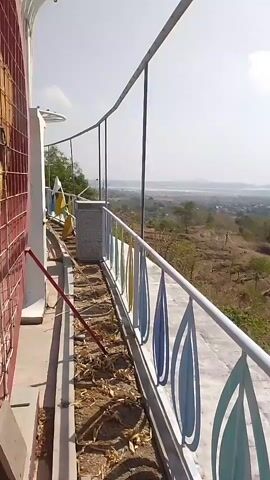The video begins with a view of a white railing with colorful leaf-shaped cutouts, curving to the right. A broom is visible on the left, leaning against the railing. Beyond the railing, a landscape unfolds with dry, brown terrain dotted with sparse trees and some buildings in the distance. The sky is bright and hazy. As the video progresses, the camera pans to the right, revealing more of the dry, hilly landscape. Terraced fields are visible on the slopes, and a plume of smoke rises from a distant area. Several paths or roads wind through the terrain. The railing continues to be a prominent element throughout the video, marking the edge of what appears to be an elevated viewing area or walkway. The environment is arid and sunny, with a sense of open space and a somewhat barren natural setting. There are no people visible in the video.