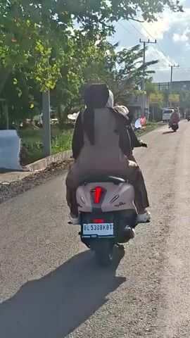 A motorcycle with two riders is traveling down a paved road. The rider in the back is wearing a brown headscarf and matching brown clothing. The person in front is wearing a black headscarf and similar brown clothing. The motorcycle is a light brown color and has a license plate that reads BL 5308 KBT. The road is bordered by grass and trees. The sun is casting shadows on the road, indicating it is daytime.