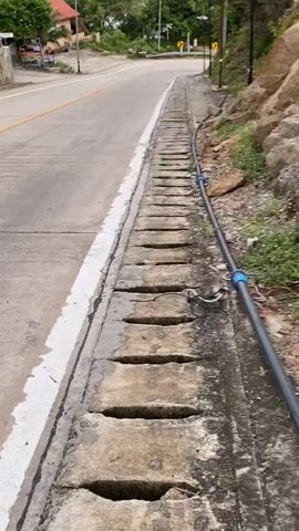 The video presents a scene from a low vantage point looking down an asphalt road with a painted median and white edge line. A concrete drainage ditch, covered with slotted grates, runs along the roadside. A black pipe with blue couplings follows the length of the ditch. On the right side of the road, a rocky hillside is covered with vegetation. Streetlights line the road. In the distance, the view opens to an ocean with a cloudy sky overhead.  Buildings are visible on the left side of the road.
