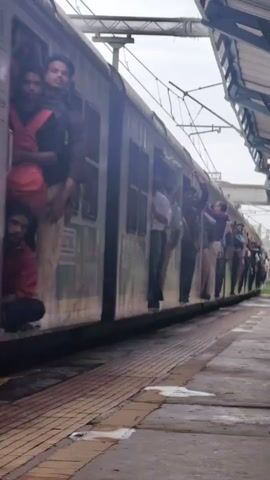 A crowded train is seen from the side at a station. Several people are leaning out of the open doors, some holding onto the doorframes. The train appears to be moving or has just arrived at the station, as indicated by the blurred motion of the train and the passengers. The platform is visible in the foreground, with yellow tactile paving along the edge. The sky is overcast.