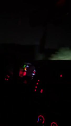 The video shows a first-person perspective from inside a car at night. The dashboard lights are illuminated, with red and white lights visible. The steering wheel is also visible. Outside the car, the scene is dark with faint outlines of trees and a road. The car appears to be moving, as suggested by the changing lights on the dashboard and the motion blur of the exterior. The overall impression is of driving through a dark, possibly rural, environment at night.