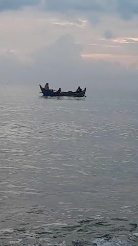 A boat containing three people moves across calm water. The sky is cloudy and gray. The foreground shows small, light-colored waves moving towards the camera. The boat is visible in the distance, and the figures appear to be rowing and steering. The overall atmosphere is quiet and still.