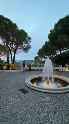 The video opens with a wide shot of a courtyard with a circular fountain in the center. The fountain has water spraying upwards and is illuminated by soft lighting. The courtyard is paved with cobblestones, and there are trees and some bushes around the perimeter. In the background, there is a glimpse of a cityscape or landscape, suggesting the location is elevated. A woman in a red dress is visible in the distance, standing near the edge of the courtyard. The sky is a pale blue, indicating it's either early morning or late evening. The camera then pans and zooms to a slightly blurry scene showing people and cars on a driveway. Some individuals appear to be interacting near a car. The camera then shifts to a close-up of a young man's face as he looks towards the camera, appearing surprised or concerned. He is wearing a white shirt and several rings. In the background, another woman is partially visible. The setting appears to be outdoors, possibly in front of a building with classical architectural elements, like columns. The overall impression is a transition from a serene outdoor environment to a more dynamic social interaction.