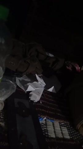 The video shows a dimly lit space. The lighting is poor, making details difficult to discern. Visible objects include crumpled pieces of white paper, a brown garment, a plastic bottle with a green label, a plastic bag, and a dark rectangular object with a design. These items are placed on a surface with a woven or patterned texture. The overall impression is one of clutter in a dark environment.