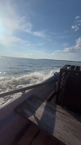 This video captures a boat ride on a body of water, likely a river or lake, under a bright, sunny sky. The perspective is from within the boat, looking forward and slightly to the side. In the foreground, we see wooden planks and parts of the boat's interior, including a metal railing. A large, weathered metal cage or crate is prominently positioned on the boat.

The boat is moving, creating a wake of white water that trails behind it. The water itself is a deep blue, reflecting the clear sky dotted with fluffy white clouds. The sun is shining brightly, casting a shimmering reflection on the water's surface. In the distance, a tree-lined shore is visible, suggesting a rural or natural setting. The overall atmosphere is peaceful and serene, conveying a sense of leisurely travel on a beautiful day. There is no visible text in the video. The time of day appears to be late morning or early afternoon, given the position of the sun. The weather is fair and pleasant. The location context provided is Вурманкасы, Russia, which aligns with the natural scenery depicted.