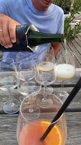 A person wearing a blue t-shirt is pouring sparkling wine from a green bottle into several wine glasses on a wooden table. One glass in the foreground contains a pinkish drink with an orange slice and a black straw. The glasses are arranged in a semi-circle, with some already containing a small amount of sparkling wine. In the background, there is greenery and a glimpse of a car.
