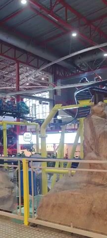 The video shows an indoor roller coaster running its course. The roller coaster train is blue and green with multiple rows of passengers, all of whom appear to be children or young adults. The roller coaster track is yellow and supported by a red metal framework visible in the background, along with exposed white curved pipes and overhead lighting. In the foreground, there is a faux rock formation and a blue metal railing with a wooden top. A sign that says "I'm Good" is visible in the background. The overall environment appears to be a brightly lit indoor amusement park or entertainment center.