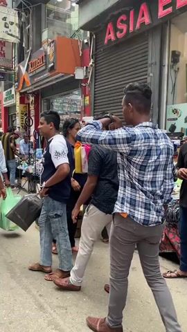 The video shows a bustling street scene with numerous people walking by. The foreground features a man in a blue and white plaid shirt looking to his left. Behind him, a man in a dark shirt and light-colored pants walks away from the camera, and another man in a dark blue shirt with a white design on the sleeve, and blue jeans, also walks away from the camera carrying a black plastic bag. In the background, there are several shops with signs displaying various products and services, including "Xmart," "Mobile," and "REPAIRING." People are seen entering and exiting the shops, and some are carrying shopping bags. The overall impression is one of a busy commercial area with high foot traffic.