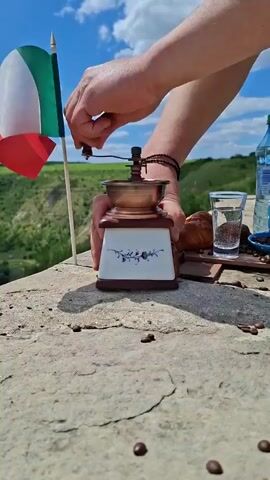 A person's hands are grinding coffee beans using a vintage manual coffee grinder. The grinder is placed on a stone surface, with a small Italian flag in the background. Next to the grinder are a glass of water, a croissant, a cup, and a bottle of water. The scene appears to be outdoors with a scenic view of hills and a blue sky with clouds.