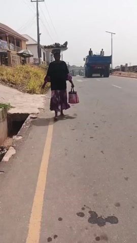 A woman walks along a paved road, carrying something on her head in a tray and a pink bag in her left hand. She is wearing a black top and a purple, floral skirt. A blue truck with two people standing in the bed is visible in the distance on the same road. Buildings and foliage line the side of the road. There are what appear to be oil stains on the road. The sky is overcast. The camera follows the woman, eventually focusing on the road.