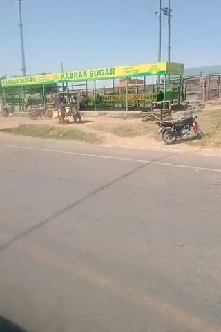 The video shows a roadside scene in what appears to be a rural area. The road is paved and a few vehicles, including a minibus and several motorcycles, are seen.  A structure advertising "Kabras Sugar" is visible on the side of the road. People are walking and standing in the vicinity, near shops and businesses, which include "Al Bahrain." The weather appears sunny and the sky is clear.