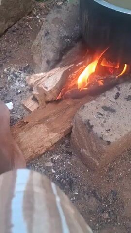 The video shows a fire burning under a large pot, possibly for cooking outdoors. The fire is built using wood and bricks. A person is sitting near the fire, with their foot visible in the foreground. The foot has white nail polish. The surrounding area appears to be a dirt or ground surface with some rocks and debris.
