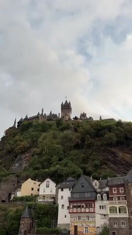 A video taken from a low angle looking up at a large, historic castle perched on a lush green hillside. Several buildings, appearing to be homes or shops, are clustered at the base of the hill, in front of the castle. The sky above is partly cloudy, with streaks of blue visible between the white clouds. The video is static, with no movement or discernible action taking place.