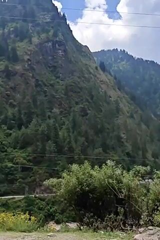 The video captures a scenic drive through the mountainous region of Manikaran, India. The landscape is dominated by steep, lush green mountains, densely covered with coniferous trees.

The initial shot (0:00-0:01) focuses on a tall, forested mountain slope under a cloudy sky. Power lines are visible in the upper frame.

From 0:01 onwards, the camera reveals a winding paved road, suggesting the perspective from a moving vehicle. On the left side of the road, several multi-story buildings and houses are nestled into the mountain slopes, forming a small settlement. Further along, more distant mountain ranges appear, partially obscured by clouds or atmospheric haze. Power lines stretch across the sky, crisscrossing the valley. The foreground features rocks, shrubs, and unpaved areas alongside the road. The sky remains overcast with prominent gray clouds throughout the video.

The scene depicts a blend of rugged natural beauty with signs of human presence in the form of roads, buildings, and infrastructure. No people are visible in the video. A white sign is briefly visible on the left side of the road around 0:02, but its text is unreadable. The overall atmosphere is serene and majestic, showcasing the characteristic topography of the Himalayas.
