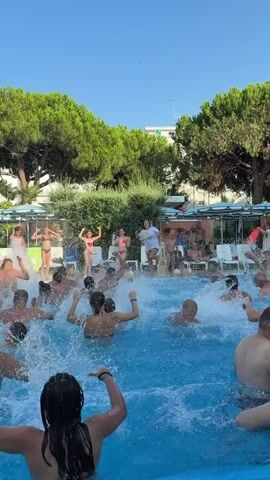 A group of people are enjoying themselves in a swimming pool. The water is splashing, indicating active movement and play. Several individuals are visible in the foreground and mid-ground, with some in the background. Trees and lounge chairs are present around the pool area, suggesting an outdoor recreational setting, possibly a resort or hotel. The sky is clear and blue, indicating pleasant weather. Some people on the edge of the pool are observing the activity. One person is standing and appears to be leading some sort of activity or dance.