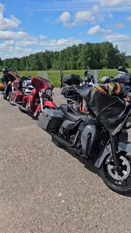 The video shows a row of motorcycles parked in a paved lot on a sunny day with scattered clouds. The first motorcycle is a gray touring bike with a black leather jacket draped over it. The other motorcycles include models in red, blue, and silver. A green field with a line of trees is visible in the background. At the end of the row, a couple of people are standing.