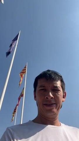 The video shows a man standing outside against a clear blue sky with several flags in the background. He is wearing a white t-shirt and sunglasses. The man smiles at the camera, then puts on his sunglasses. The flags appear to be the European Union flag, the Catalan flag, and the Spanish flag. The video is shot from a low angle, looking up at the man and the flags.