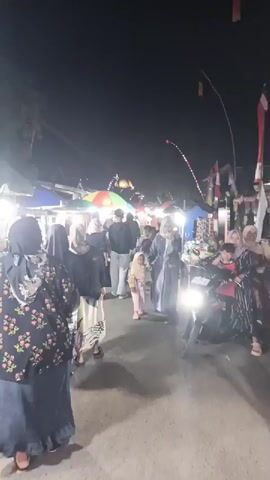A nighttime outdoor market scene shows a diverse group of people, some walking, others standing or riding a motorcycle. Multiple stalls line the pathway, with bright lights illuminating the merchandise. In the foreground, a woman in a floral top and dark skirt walks away from the camera. To her right, a child in a light pink outfit and headscarf walks with an adult. A motorcycle with its headlight on passes by in the foreground. In the background, more people are seen browsing the stalls, and a colorful umbrella stands out. The sky is dark, indicating it is night.