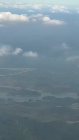 The video displays an aerial view of a landscape from above, likely from an airplane. The upper portion of the frame shows a blue sky with scattered white cumulus clouds. Below the clouds, a layer of atmospheric haze obscures the distant horizon. The mid-ground and foreground feature a complex network of interconnected water bodies, appearing as numerous lakes or reservoirs with irregular shorelines. These bodies of water are surrounded by rolling hills and landmasses densely covered in dark green or grey foliage, forming multiple islands and peninsulas. In the far distance, through the haze, faint outlines of what appear to be roads or cleared areas can be discerned. The camera remains largely static, with minimal discernible movement, offering a steady panoramic shot of the terrain.