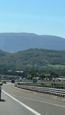 The video shows a perspective from a moving vehicle on a highway. The road curves to the left, with a white line marking the lane. A metal guardrail runs alongside the road. In the distance, there are multiple lanes of traffic, with various vehicles including cars and trucks. Beyond the highway, there are buildings and industrial structures, as well as numerous utility poles and electrical lines. Further in the background, a lush green, tree-covered hillside rises steeply, and behind that, a range of hazy blue mountains stretches across the horizon under a clear blue sky. The lighting suggests it is daytime with bright sunlight.