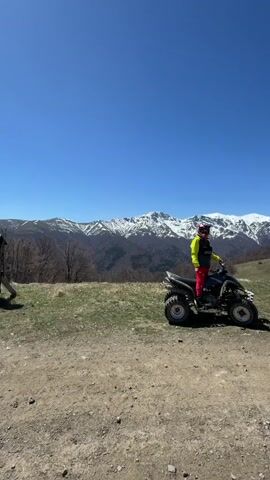 The video shows a person sitting on an all-terrain vehicle (ATV) on a dirt path in a mountainous region. The person is wearing a yellow and black jacket, red pants, and a helmet. The ATV is dark-colored. In the background are snow-capped mountains under a clear blue sky. The terrain appears to be grassy and somewhat barren with scattered trees. The camera is static and the ATV is not moving.