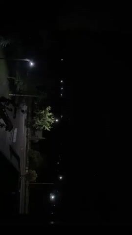 The video displays a nighttime scene along a street lined with trees and lampposts. A faint purple light illuminates the sky in the distance, suggesting distant lightning. The street itself is dark, with only the lampposts providing illumination. No people or vehicles are visible in the immediate frame.