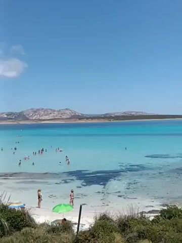 The video shows a wide view of a crowded beach on a sunny day. The water is a clear turquoise, with many people swimming and wading in the shallow areas. The beach itself is sandy, with numerous umbrellas and sunbathers dotting the shoreline. In the distance, a landmass with hills is visible across the water. The sky is a vibrant blue with a few wispy white clouds. The camera pans slightly, revealing more of the beach and the expanse of the ocean.