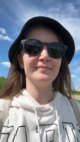 A woman wearing a bucket hat and sunglasses looks into the camera and smiles slightly, then looks to her right. She is wearing a white hoodie and has a backpack. The background shows a blue sky with some clouds and a building in the distance. The camera is held at chest level.