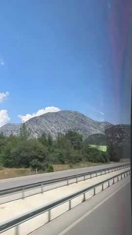 The video shows a view from a moving vehicle, likely a bus, looking out a window. The scene is dominated by a large, rugged, rocky mountain under a bright blue sky with scattered white clouds. In the foreground, there is a multi-lane highway with guardrails. Trees and vegetation line the side of the road, with more trees on the lower slopes of the mountain. The lighting suggests it is daytime, likely sunny. The movement of the vehicle causes a slight blur and a sense of motion.