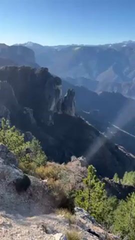 The video shows a scenic landscape with mountains and trees under a clear blue sky. The camera pans slowly to reveal a vast canyon or valley with rocky cliffs and sparse vegetation. Some distant peaks are covered in snow. The environment appears rugged and natural, with no visible signs of human development in the valley below. The foreground features more vegetation and a rocky edge.