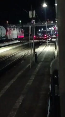 The video shows a train station at night. Several trains are visible on the tracks, illuminated by headlights and station lights. The station platform is paved and marked with white lines. There are trash cans and light poles along the platform. The camera appears to be positioned inside a train, looking out at the station. The lighting is dim, with strong shadows cast by the train and station lights.
