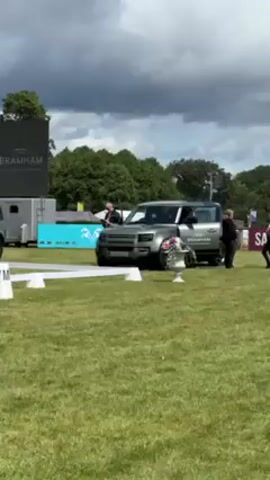 The video shows a parking area with two Land Rover Defenders parked on the right side. A person in a blue suit is standing next to the first Land Rover, seemingly cleaning it. 


Behind the Defenders, there is a large screen displaying "DEFENDER" and "BRAMHAM" logos, indicating a promotional event. To the left, there is a sound system, likely for event announcements or music. 


The environment is a grassy field, possibly an event space, marked with white lines and cones, potentially outlining a parking area or an obstacle course. The sky is partly cloudy, suggesting a fair-weather day. 


Overall, the scene appears to be from a promotional event or car show for Land Rover Defender.