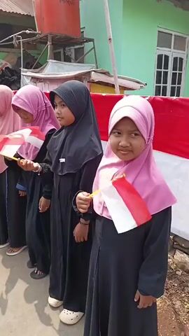 A group of young girls, dressed in hijabs and dark colored dresses, stand in a line, each holding a small Indonesian flag with a yellow pole. They are positioned outdoors on a paved surface, possibly a street or walkway. Behind them, a red and white banner is displayed, possibly representing the Indonesian flag. In the background, there are buildings and some outdoor structures, suggesting a residential or community area. The girls appear to be participating in a patriotic event or celebration. The lighting suggests it is daytime.