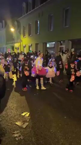 At night, a crowd of people are moving along a street with green buildings on one side. Many of the people are wearing colorful costumes, including tutus and glow sticks. Some are pulling wagons. There is a drum sound in the background. The people are walking towards the camera. The scene is illuminated by streetlights and the lights from the costumes.