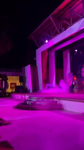 A performance is underway on an outdoor stage at night, illuminated by pink and green lights. Several performers are present. On the left, a person dressed as Beetlejuice, in a black and white striped suit with a red tie and white face paint, stands with arms outstretched. To the right of Beetlejuice, a person in a black dress and dark hair stands near a barrel that is emitting green smoke. In the background, other figures can be seen on stage. The stage has multiple levels and is decorated with props, including barrels and what appear to be coffin-like structures. The audience area in front of the stage is also lit with pink lights. Smoke is present on the stage, creating a dramatic effect.