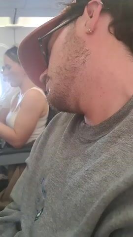 The video shows a man wearing a gray sweater, glasses, and a baseball cap sitting in an airplane seat. He appears to be resting or sleeping. In the background, a woman with headphones is sitting in the seat in front of him. The interior of the airplane is visible, with overhead compartments and other passengers.