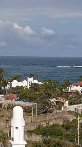 The video shows a scenic coastal view. It starts with a panorama of several white and orange buildings, including a distinct white dome. Palm trees are interspersed among the buildings. The scene then shifts to focus on the blue ocean, where waves are visible breaking in the distance. A few ships can be seen on the horizon. The sky is predominantly blue, with scattered white clouds.