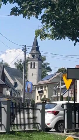 A white car is parked near a fence. A church steeple is visible behind buildings. Two people are riding a bicycle.