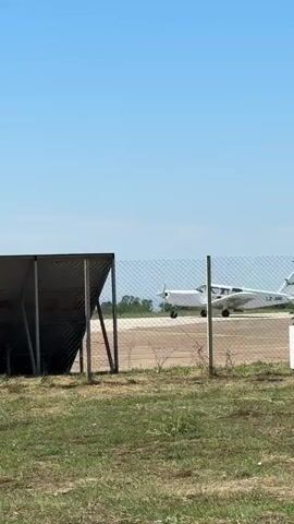 A small white airplane with LZ-ANI visible on its tail sits on a tarmac runway, behind a chain-link fence, under a clear blue sky. In the foreground, grass and dirt cover the ground. To the left, a metal structure with a red frame and a dark panel is partially visible.