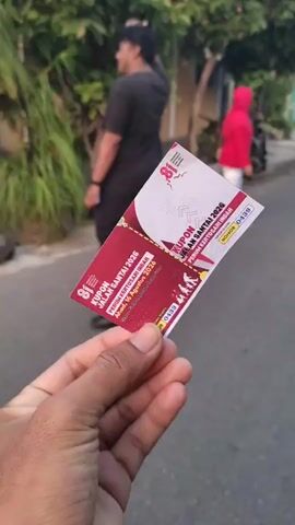 The video begins with a close-up shot of a hand holding a red and white coupon. The coupon has text in Indonesian that appears to be related to an event. In the background, a person wearing a black shirt and pants is walking away from the camera, and another person in a red hijab and white pants is visible further down a street. The environment appears to be a residential area with trees and buildings. As the video progresses, the camera pans slightly, revealing more of the street scene, including more people, a motorcycle, and what looks like stalls or decorations for an event. The coupon remains the central focus throughout.
