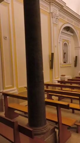 The video opens with a close-up of a dark, cylindrical pillar in the foreground. The camera then pans to the right, revealing the interior of a church. Rows of empty wooden pews with red backs and legs fill the space, arranged on either side of a central aisle. The walls are painted a pale yellow with white decorative molding and architectural details. To the right, an altar is visible, adorned with candlesticks and religious iconography. A statue of the Virgin Mary holding the infant Jesus stands prominently on a raised platform near the altar. Further down the aisle, on the right side, another niche in the wall contains a statue, possibly of a saint, lit by a spotlight. A stained-glass window with a circular design is visible on the ceiling arch above the altar. The overall lighting is dim, suggesting an evening or interior setting, with spotlights illuminating specific areas like the statues and the altar. The floor is tiled in a pattern of light and dark colors. The camera continues to pan, showing more of the pews and the church's architectural features, before the video ends.