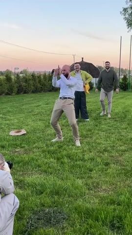 Here's a description of the video:

The video shows a man dancing on a grassy lawn. He's bald, has a beard, and is wearing a white dress shirt and khaki pants. He’s dancing with enthusiasm, making various gestures and movements, seemingly for the camera. In the background, two other men are watching and smiling. One of them is wearing a light green jacket over a white shirt and dark pants. The other is wearing a green long-sleeve shirt and khaki pants. A photographer in beige clothing is partially visible in the foreground, likely filming or photographing the man dancing. The background includes a fence, some trees, and a house or building in the distance under a dusky sky. There's also a hat lying on the grass near the dancing man. The audio includes music.