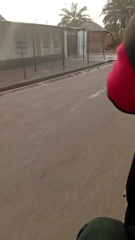 The video shows a street in a small town with modest houses and palm trees. The camera is moving, likely mounted on a motorcycle or bicycle, filming from behind. At least two other motorcycles pass. People are visible on the side of the road.