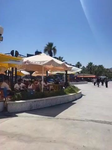 The video begins with a view of an outdoor cafe with umbrellas providing shade for patrons. Palm trees and buildings can be seen in the background, along with a clear blue sky. As the video progresses, the camera pans to show a promenade with people walking. A large cruise ship is visible in the distance, out on the water. The cruise ship appears to be docked or maneuvering in a harbor. The scene is bright and sunny, suggesting a warm day. There are also some trash receptacles and a lamppost visible along the promenade. The video ends with a close-up view of the cruise ship against the backdrop of the ocean and a distant, hilly coastline.